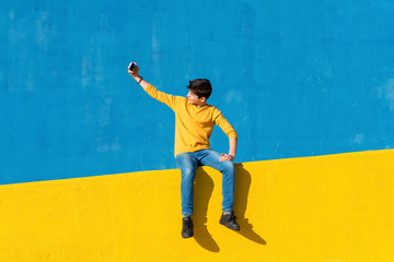 Front view of a young boy wearing casual clothes sitting on a yellow fence against a blue wall while taking a selfie with a smartphone