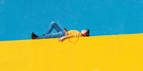 Front view of a young boy wearing casual clothes lying on a yellow fence against a blue wall while...