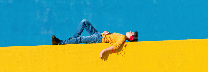 Front view of a young boy wearing casual clothes lying on a yellow fence against a blue wall while using a mobile phone to listening music by headphones - Powered by Adobe