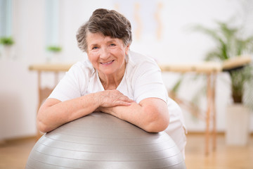 Senior woman leaning on grey gymnastic ball at physiotherapy center