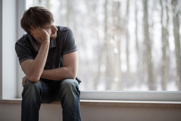 Thoughtful young man sitting on windowsill looking through the window