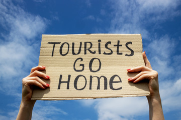 Text Tourists go home on a cardboard plate in hands. Blue sky with clouds background. Stop mass...