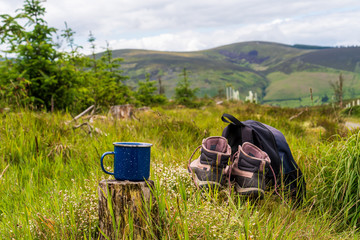 Selective focus on a camping blue enamel mug with hot coffee on a tree stump next to a pair of...