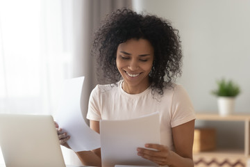 Happy african woman sit at table reading paper checking bills