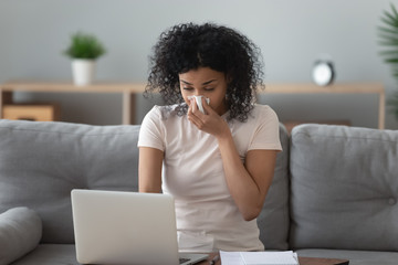 Allergic african woman blowing nose in napkin sit at home