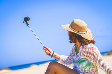 Fototapeta premium Trendy tourist doing a video with action cam on an amazing tropical beach during vacation Beautiful woman with straw hat make social story sitting on white sand Travel and new trend technology concept