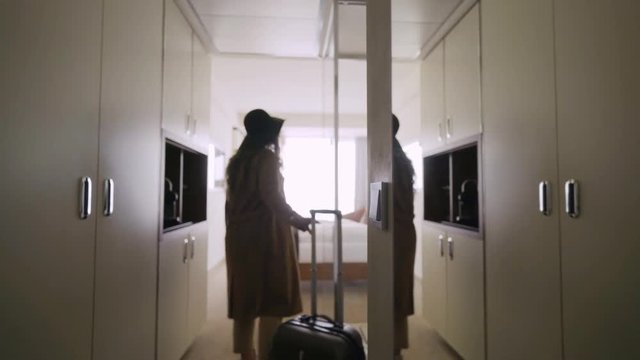 Back view woman entering in hotel apartment. Faceless female in hat with suitcase moving inside room bright illuminated daylight from full length window. Concept comfortable vacation