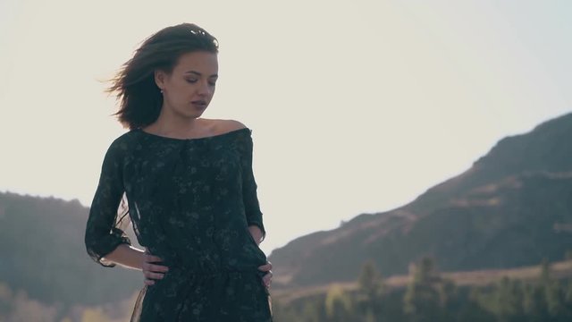 beautiful short haired girl in dark dress with loose sleeves poses against old mountains on windy spring day