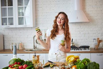 Young woman drinking green smoothie and fresh water with cucumber, lemon, leaves of mint on the kitchen table with fruits and vegetables. Healthy eating concept. Vegan meal and detox menu
