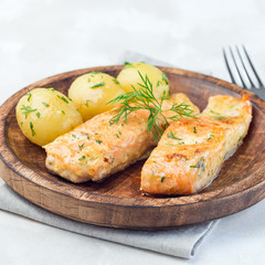 Baked salmon in creamy sauce with young boiled  potato topped with melted butter and chopped dill on a wooden plate, square format