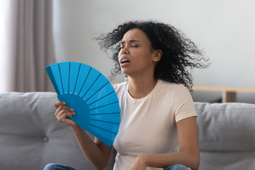 Overheated african young woman feeling hot waving fan at home