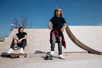 Father and his little son dressed in the casual clothes ride skateboards and have fun in a skate park with slides outside at the sunny day