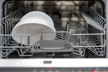 Opened modern dishwasher machine with plates and cups on kitchen