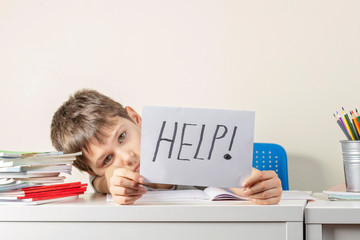 Sad tired frustrated boy sitting at the table with many books and holding paper with word Help....