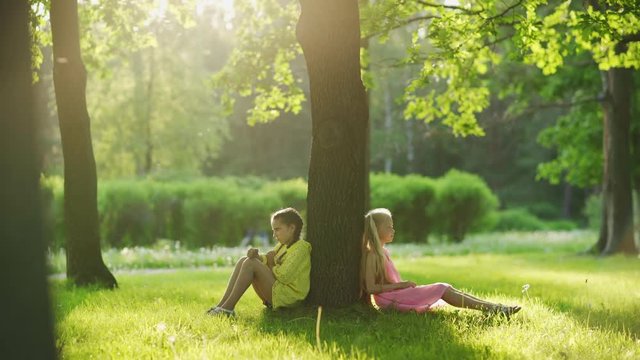Tracking right shot of two unhappy little girls sitting on grass in park, leaning on tree trunk back to back and sulking after best friends conflict on sunny summer day