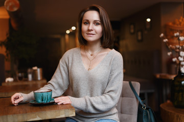 Young Caucasian European woman sitting on a chair at the cafe with cup of tea in casual sweater and jeans in cozy interior