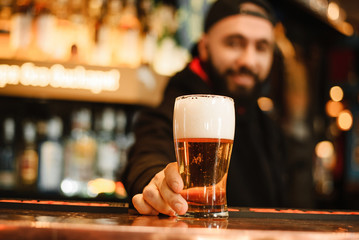 Bearded and smiling barman gives beer. Cool and courageous bar. A happy barman holds a beer in his...