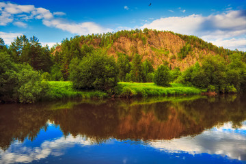 Summer landscape river with reflecting trees against the backdrop of forests and mountains.