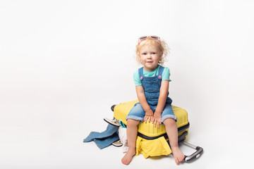 Funny curly little baby girl in a hat, T-shirt and jeans with a yellow suitcase is smiling on white background waiting for an airplane. concept trip with kids.