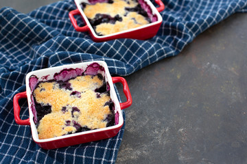 Two freshly baked blueberry cobblers in red pans on blue kitchen towel with black textured background.