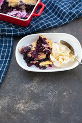 Freshly baked blueberry cobblers in red ceramic pans; some isolated on white plate in front with vanilla ice cream; blue kitchen towel; black textured bakcground.