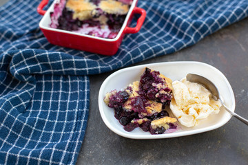 Freshly baked blueberry cobblers in red ceramic pans; some isolated on white plate in front with vanilla ice cream; blue kitchen towel; black textured bakcground.
