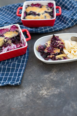 Freshly baked blueberry cobblers in red ceramic pans; some isolated on white plate in front with vanilla ice cream; blue kitchen towel; black textured bakcground.