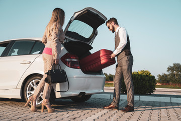 Handsome elegant man putting woman travel bag in modern car 