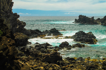 Rocky coastline near the Kai Tanata cave on Easter Island, showcasing striking black volcanic rocks, turquoise ocean waves crashing on the shore, and dramatic coastal scenery under a sunny sky.