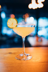 Refreshing fresh classic lime margarita cocktail in margarita glass with lemon on wooden table in front of window. Vintage retro bar interior design.
