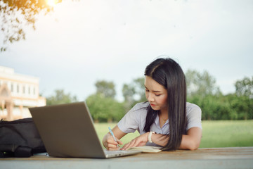 A young female university student writes a note and assignment with a laptop beside outside the campus with blure green tree background. Students self learning outside the campus.