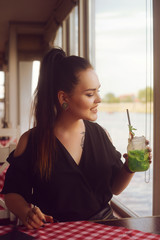 The girl in a restaurant on the ship near the river drinks mojito. Brunette with tattoo looks out...