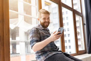 Fototapeta premium Cute young positive guy hipster with a beard is sitting on the windowsill by the big window and talking on the video communication using a smartphone and wireless high-speed Internet.