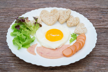 Breakfast consists of fried egg, ham, sausage, whole wheat bread, fresh vegetables, put plates on old wooden tables.