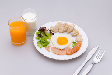 Breakfast consists of fried egg, ham, sausage, milk, orange, whole wheat bread shaped like a heart, fresh vegetables, put plates on white background