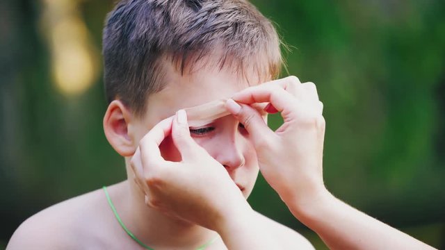 Sad boy with injury on his eyebrow. Woman's hands putting small white adhesive plaster on a wound outdoors. Close-up.