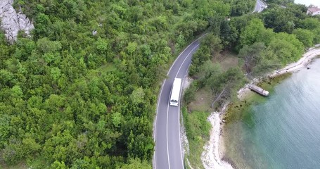 The quadcopter is flying over a narrow asphalt road running along the sea shore. We can see the turquoise sea and the seashore covered with green trees on the other side of the road