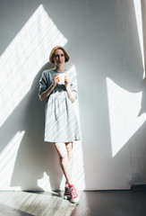 Young woman in simple grey dress with yellow cup standing against wall, full-length portrait, hard light