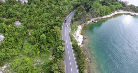 The quadcopter is flying over a narrow asphalt road running along the sea shore. We can see the turquoise sea and the seashore covered with green trees on the other side of the road