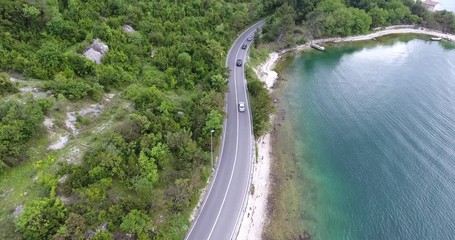The quadcopter is flying over a narrow asphalt road running along the sea shore. We can see the turquoise sea and the seashore covered with green trees on the other side of the road