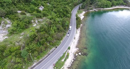 The quadcopter is flying over a narrow asphalt road running along the sea shore. We can see the turquoise sea and the seashore covered with green trees on the other side of the road