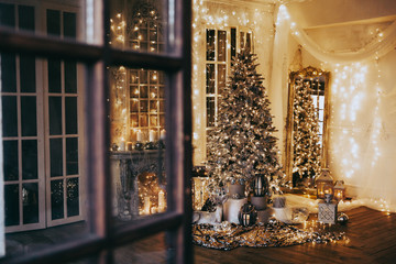 warm cozy evening in Christmas room interior design,Xmas tree decorated by lights gifts,toys, deer,candles, lanterns, garland lighting indoors fireplace.holiday.magic New year
