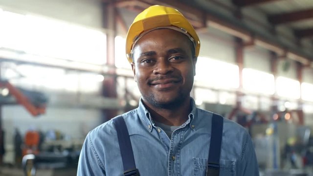 Pan close-up backlit shot of black smiling technician looking relaxed and confident, gazing at camera, with bright sunny factory hall and large windows flooded with daylight in blurred background