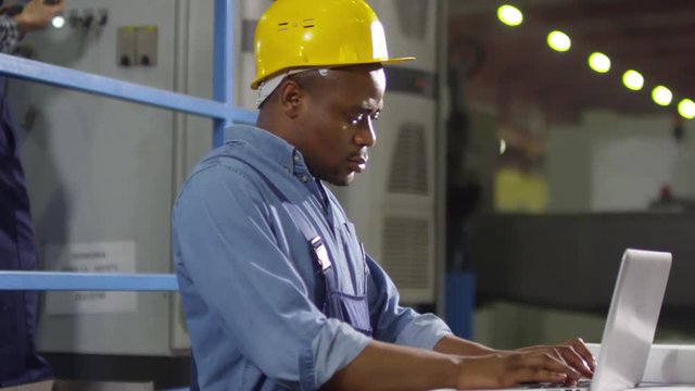 Panning waist-up shot of thoughtful Afro-American technician, in overalls and hardhat, writing shift report on laptop, with automated production line equipment of modern factory in background