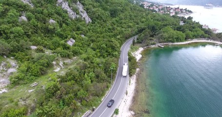 The quadcopter is flying over a narrow asphalt road running along the sea shore. We can see the turquoise sea and the seashore covered with green trees on the other side of the road