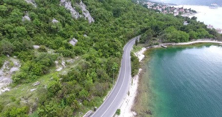 The quadcopter is flying over a narrow asphalt road running along the sea shore. We can see the turquoise sea and the seashore covered with green trees on the other side of the road