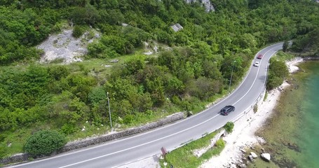 The quadcopter is flying over a narrow asphalt road running along the sea shore. We can see the turquoise sea and the seashore covered with green trees on the other side of the road