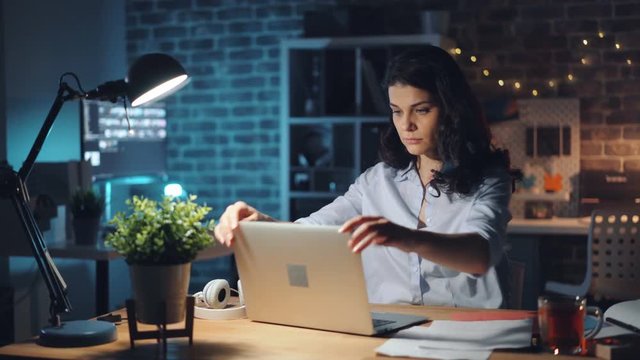 Young Woman Is Turning Off Laptop And Light In Office At Night And Leaving Work After Long Workday. Hard-working People, Overtime Job And Millennials Concept.