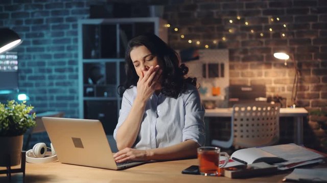 Exhausted young lady is typing with laptop then yawning working in office at night being exhausted at work. Business, hard-working people and millennials concept.