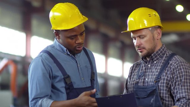 Panning medium two shot of factory workers, Afro-American and Caucasian, both wearing blue overalls and yellow hardhats discussing production plans and filling out checklist on clipboard